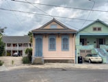 A quaint residential street featuring a small, charming house painted in pastel peach with blue accents. The house has ornate decorative trim, shutters, and a central entrance with steps. Adjacent to it is a similarly styled house painted in mint green. In the background, there is a two-story building with a darker exterior and white balconies. Overhead, power lines crisscross against a cloudy sky.