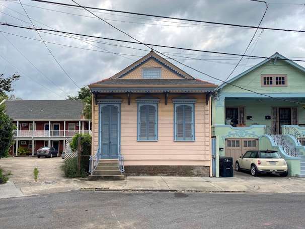 A quaint residential street featuring a small, charming house painted in pastel peach with blue accents. The house has ornate decorative trim, shutters, and a central entrance with steps. Adjacent to it is a similarly styled house painted in mint green. In the background, there is a two-story building with a darker exterior and white balconies. Overhead, power lines crisscross against a cloudy sky.