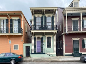 A row of three colorful two-story houses with distinct New Orleans-style architecture featuring ornate ironwork on the balconies. The center house is painted light green with dark green shutters and a purple door. The house on the left is orange, and the one on the right is maroon, both with dark-colored shutters and similarly styled balconies. A green car is parked on the street in front, and utility lines cross the upper portion of the image.