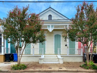 A charming wooden house with pale green shutters and a decorative gable, flanked by two flowering trees with red blossoms. The building features intricate white trim and a small front porch with stairs leading up to it. Recycling bins are visible on the left side of the house, and there are additional colorful houses in the vicinity.