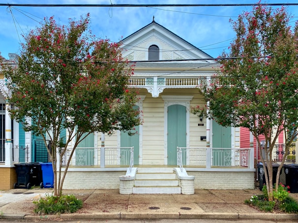 A charming wooden house with pale green shutters and a decorative gable, flanked by two flowering trees with red blossoms. The building features intricate white trim and a small front porch with stairs leading up to it. Recycling bins are visible on the left side of the house, and there are additional colorful houses in the vicinity.