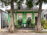 A vibrant, green-painted house with purple accents is flanked by two large trees in the foreground. The house has decorative elements like window trims painted in orange and white, lush greenery with tall plants at the front, and ornamental masks mounted on the wall. A small, white sculpture is centered among the plants.
