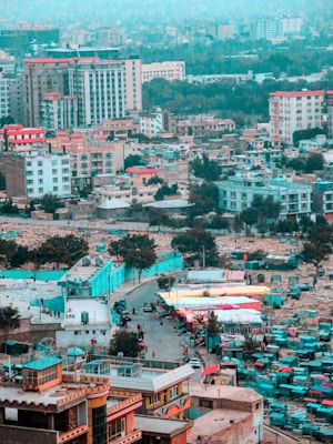 A bustling cityscape featuring numerous buildings with varied architecture, including residential and commercial high-rises. The streets are lively, with people and vehicles moving through a marketplace area lined with colorful tents.