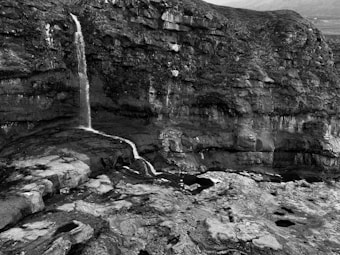 A dramatic waterfall cascades down a rugged cliff face, set against a backdrop of textured rock formations. The landscape is monochrome, highlighting the intricate details of the stone and the flow of water. The environment appears desolate yet majestic, with a sense of natural rawness and untouched beauty.