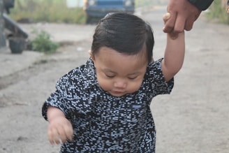 A young child with dark hair and wearing a patterned black and white shirt is holding an adult's hand. The child appears to be taking a step forward on a dirt path. In the background, there is a blurred blue vehicle and some greenery.