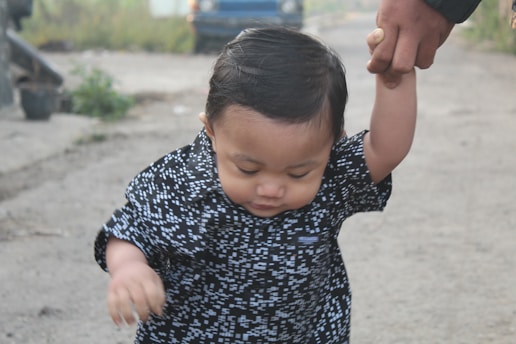 A young child with dark hair and wearing a patterned black and white shirt is holding an adult's hand. The child appears to be taking a step forward on a dirt path. In the background, there is a blurred blue vehicle and some greenery.