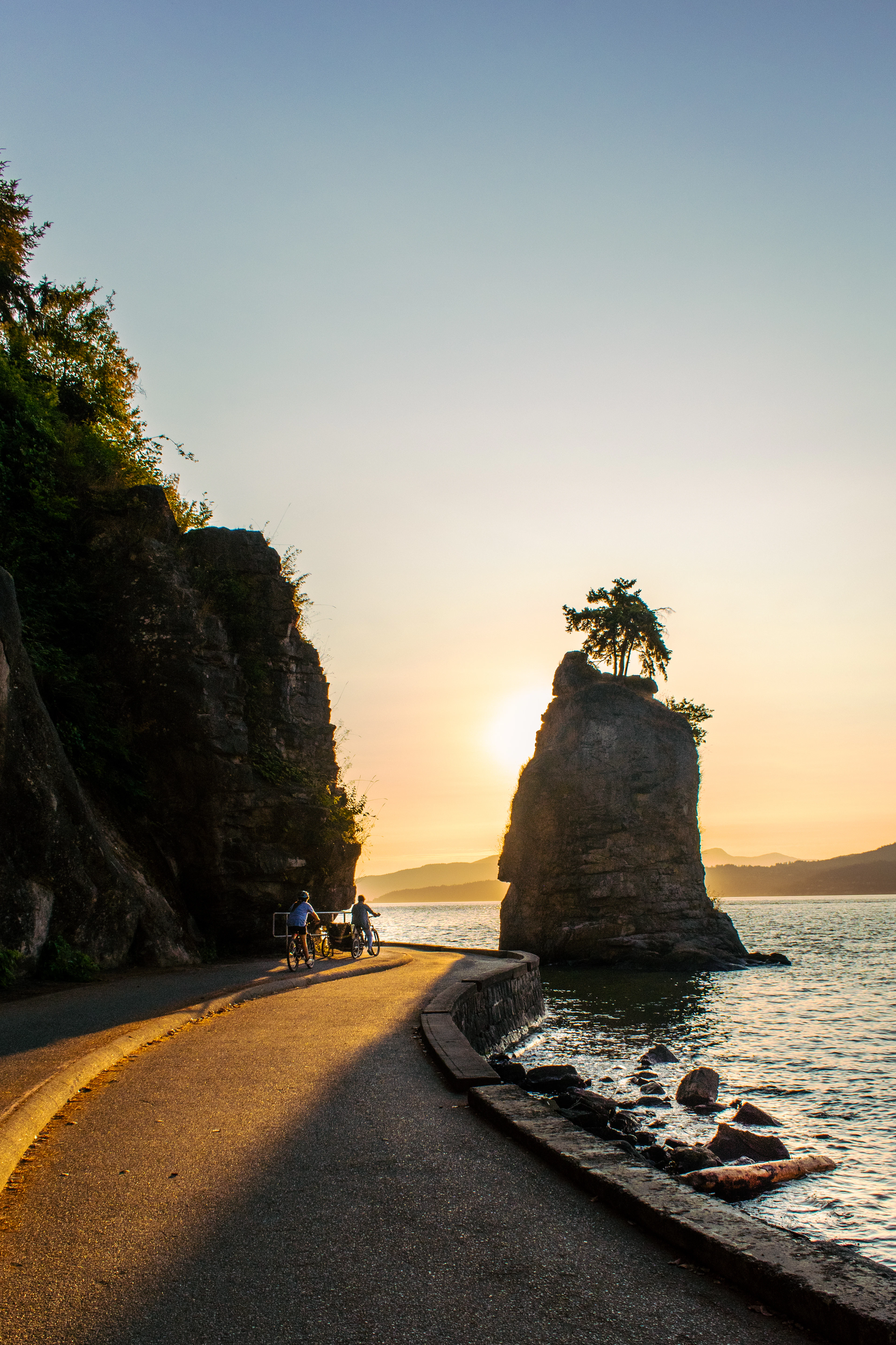 Cyclists on the Stanley Park seawall past Siwash Rock at sunset
