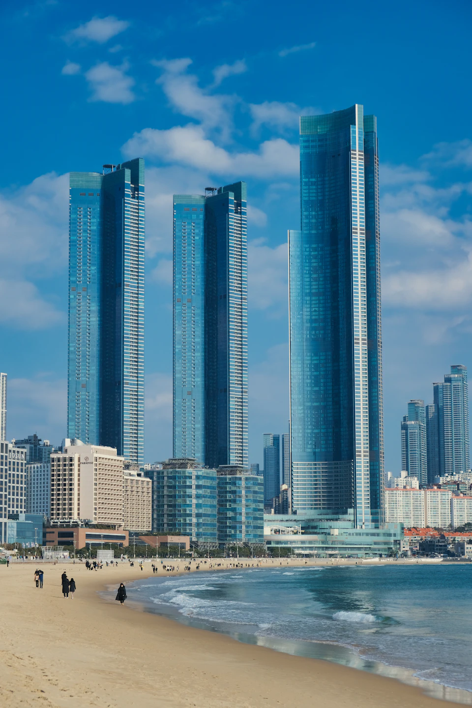 The image depicts a bustling cityscape with three tall buildings, each adorned with blue glass facades that reflect the clear sky above. The tallest building is situated on the right side of the frame, while the other two are positioned more centrally in the scene. In front of these structures, there's a sandy beach where several people can be seen walking along it.

The cityscape features various buildings of different heights and designs, creating an urban environment that contrasts with the natural beauty of the beach. The clear blue sky above adds to the overall aesthetic appeal of the image.