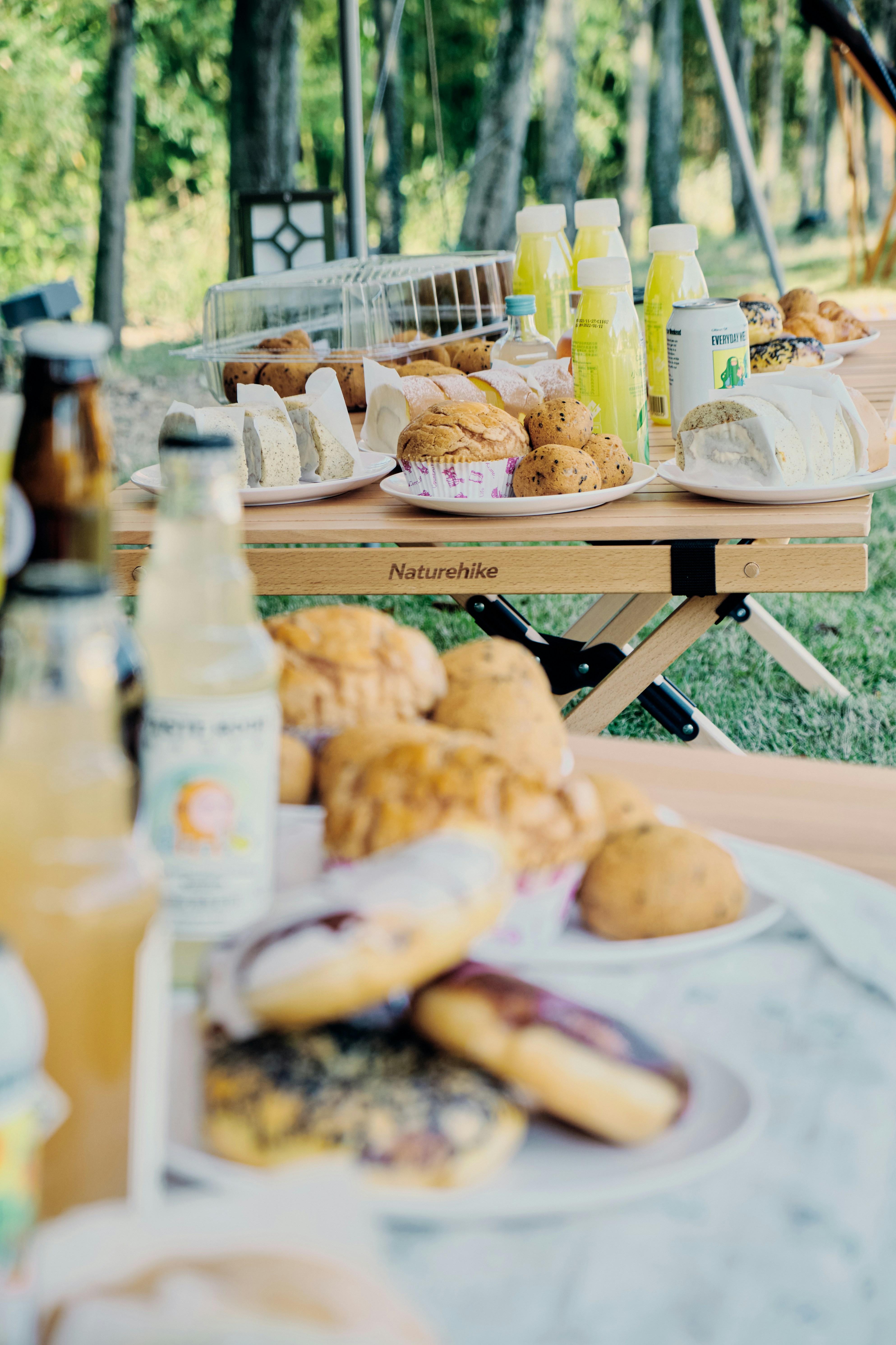 Photograph of a park picnic spread featuring loaves, buns, and pastries on a wooden Naturehike table. Bottled drinks and juice cartons line the setup, while a blurred foreground pastry tray adds depth.
