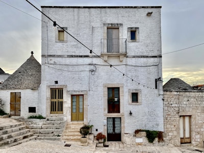 A white stone building with a rustic appearance, featuring multiple windows and a small balcony. The structure is surrounded by string lights that cross the image diagonally. On the left side, traditional conical roofs, known as trulli, are partially visible. The setting appears quiet and serene, with potted plants adorning the steps leading to the entrance.