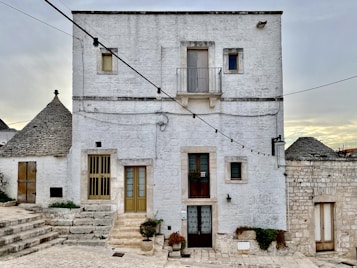 A white stone building with a rustic appearance, featuring multiple windows and a small balcony. The structure is surrounded by string lights that cross the image diagonally. On the left side, traditional conical roofs, known as trulli, are partially visible. The setting appears quiet and serene, with potted plants adorning the steps leading to the entrance.