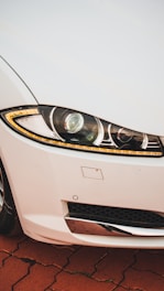 A close-up view of a car's front right headlight and part of the grille on a vehicle with a white exterior. The headlight features intricate design elements with LED strips and a reflective surface. The surrounding area includes part of the bumper and a metallic component below the light, positioned on a reddish-brick pattern ground.