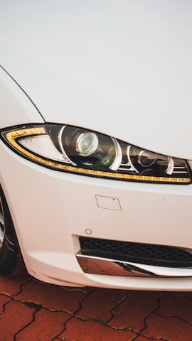 A close-up view of a car's front right headlight and part of the grille on a vehicle with a white exterior. The headlight features intricate design elements with LED strips and a reflective surface. The surrounding area includes part of the bumper and a metallic component below the light, positioned on a reddish-brick pattern ground.