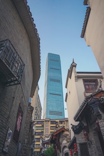 A modern skyscraper with a blue glass facade towers above traditional Chinese architecture with intricate tiled roofs. Red lanterns and brickwork details add to the cultural contrast.