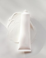 A white, cylindrical tube of cream or lotion is placed on a smooth surface. The cream is slightly smeared around the base of the tube, creating a textured and glossy appearance. The overall scene is minimalistic, with a focus on the simplicity and clean design of the product.