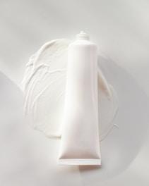 A white, cylindrical tube of cream or lotion is placed on a smooth surface. The cream is slightly smeared around the base of the tube, creating a textured and glossy appearance. The overall scene is minimalistic, with a focus on the simplicity and clean design of the product.