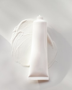 A white, cylindrical tube of cream or lotion is placed on a smooth surface. The cream is slightly smeared around the base of the tube, creating a textured and glossy appearance. The overall scene is minimalistic, with a focus on the simplicity and clean design of the product.