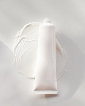 A white, cylindrical tube of cream or lotion is placed on a smooth surface. The cream is slightly smeared around the base of the tube, creating a textured and glossy appearance. The overall scene is minimalistic, with a focus on the simplicity and clean design of the product.