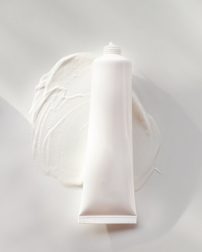 A white, cylindrical tube of cream or lotion is placed on a smooth surface. The cream is slightly smeared around the base of the tube, creating a textured and glossy appearance. The overall scene is minimalistic, with a focus on the simplicity and clean design of the product.