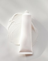 A white, cylindrical tube of cream or lotion is placed on a smooth surface. The cream is slightly smeared around the base of the tube, creating a textured and glossy appearance. The overall scene is minimalistic, with a focus on the simplicity and clean design of the product.