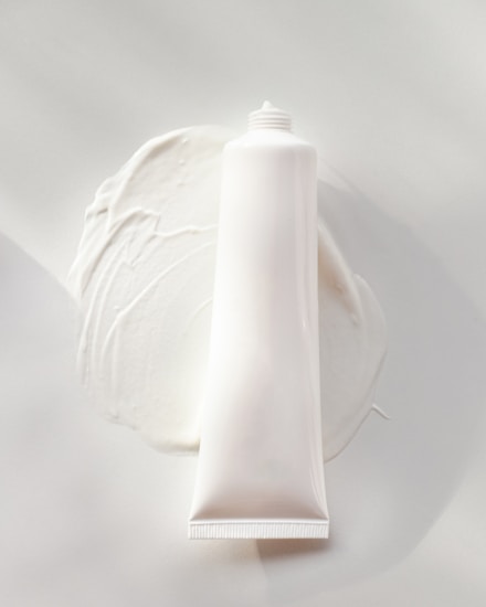 A white, cylindrical tube of cream or lotion is placed on a smooth surface. The cream is slightly smeared around the base of the tube, creating a textured and glossy appearance. The overall scene is minimalistic, with a focus on the simplicity and clean design of the product.
