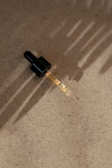A dropper with a black cap is placed on a textured, beige surface. The dropper is partially filled with a light-colored liquid. Soft, linear shadows are cast across the surface, likely from natural sunlight.