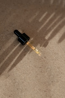 A dropper with a black cap is placed on a textured, beige surface. The dropper is partially filled with a light-colored liquid. Soft, linear shadows are cast across the surface, likely from natural sunlight.