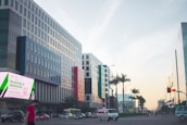 A city street lined with modern buildings on either side, featuring a prominent digital billboard advertising national ID registration. Several vehicles, including a van and cars, are visible on the road along with a person walking on a sidewalk. Palm trees are present, adding a touch of greenery against a clear sky with evening light.