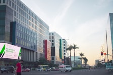 A city street lined with modern buildings on either side, featuring a prominent digital billboard advertising national ID registration. Several vehicles, including a van and cars, are visible on the road along with a person walking on a sidewalk. Palm trees are present, adding a touch of greenery against a clear sky with evening light.