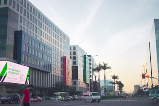 A city street lined with modern buildings on either side, featuring a prominent digital billboard advertising national ID registration. Several vehicles, including a van and cars, are visible on the road along with a person walking on a sidewalk. Palm trees are present, adding a touch of greenery against a clear sky with evening light.