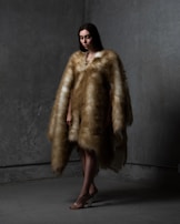 A person stands in a dimly lit space wearing a luxurious fur coat that extends to the floor. The background has a textured gray wall with subtle lighting casting shadows. The individual's posture is slightly angled, and their expression is calm, almost contemplative. Their feet are adorned with transparent high-heeled shoes.