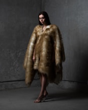 A person stands in a dimly lit space wearing a luxurious fur coat that extends to the floor. The background has a textured gray wall with subtle lighting casting shadows. The individual's posture is slightly angled, and their expression is calm, almost contemplative. Their feet are adorned with transparent high-heeled shoes.