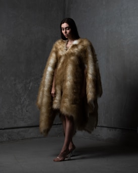 A person stands in a dimly lit space wearing a luxurious fur coat that extends to the floor. The background has a textured gray wall with subtle lighting casting shadows. The individual's posture is slightly angled, and their expression is calm, almost contemplative. Their feet are adorned with transparent high-heeled shoes.