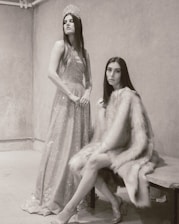 Two women are in a minimalist room with textured walls. One is standing, wearing a sparkling gown and a tiara, exuding elegance and poise. The other is seated, wrapped in a luxurious fur coat, giving a subtle yet intense gaze towards the camera.