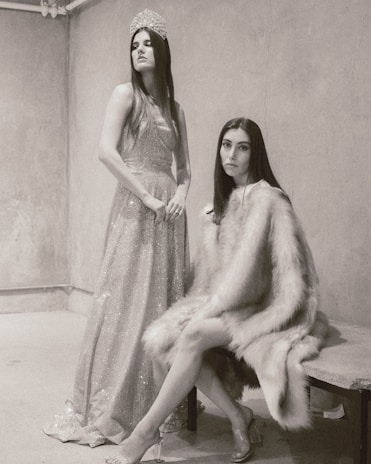 Two women are in a minimalist room with textured walls. One is standing, wearing a sparkling gown and a tiara, exuding elegance and poise. The other is seated, wrapped in a luxurious fur coat, giving a subtle yet intense gaze towards the camera.