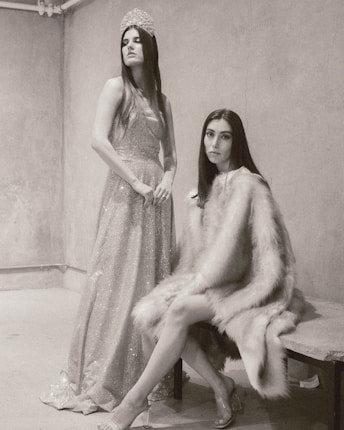 Two women are in a minimalist room with textured walls. One is standing, wearing a sparkling gown and a tiara, exuding elegance and poise. The other is seated, wrapped in a luxurious fur coat, giving a subtle yet intense gaze towards the camera.