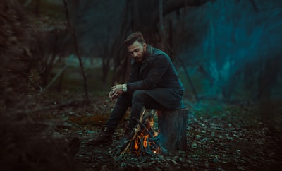 A man sits thoughtfully on a tree stump beside a small campfire in a dimly lit forest. He is dressed in dark clothing and appears contemplative, with his hands clasped together. The surrounding area is dense with trees, and the overall atmosphere is moody and mysterious.