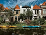 Two kayaks are moored along a small canal lined with houses. The buildings have orange-tiled roofs, and there is lush greenery along the water's edge. The sky is partly cloudy, creating a serene and picturesque setting.