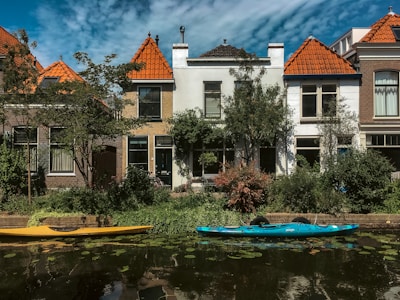 Two kayaks are moored along a small canal lined with houses. The buildings have orange-tiled roofs, and there is lush greenery along the water's edge. The sky is partly cloudy, creating a serene and picturesque setting.