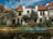 Two kayaks are moored along a small canal lined with houses. The buildings have orange-tiled roofs, and there is lush greenery along the water's edge. The sky is partly cloudy, creating a serene and picturesque setting.