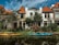 Two kayaks are moored along a small canal lined with houses. The buildings have orange-tiled roofs, and there is lush greenery along the water's edge. The sky is partly cloudy, creating a serene and picturesque setting.