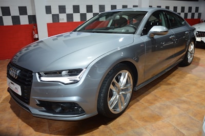 A sleek, silver luxury sedan is parked indoors on a polished tile floor. The car features a modern design with distinct headlights, a prominent front grille, and large alloy wheels. Adjacent white and red walls provide a contrasting backdrop.