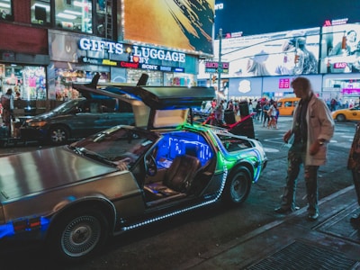 A modified DeLorean car with open gull-wing doors is parked on a bustling city street at night. The car is illuminated with bright, multicolored LED lights, drawing attention from passersby. In the background, various shops with neon and LED signage are visible, along with people walking along the sidewalk and taxis passing by. The scene has a lively and vibrant urban atmosphere.