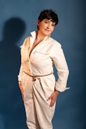 A person is posed confidently against a plain blue background. They are wearing a stylish white jumpsuit with a belt, and their hands rest on their hips. Their expression is relaxed and approachable.