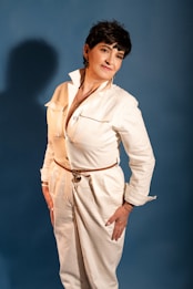 A person is posed confidently against a plain blue background. They are wearing a stylish white jumpsuit with a belt, and their hands rest on their hips. Their expression is relaxed and approachable.