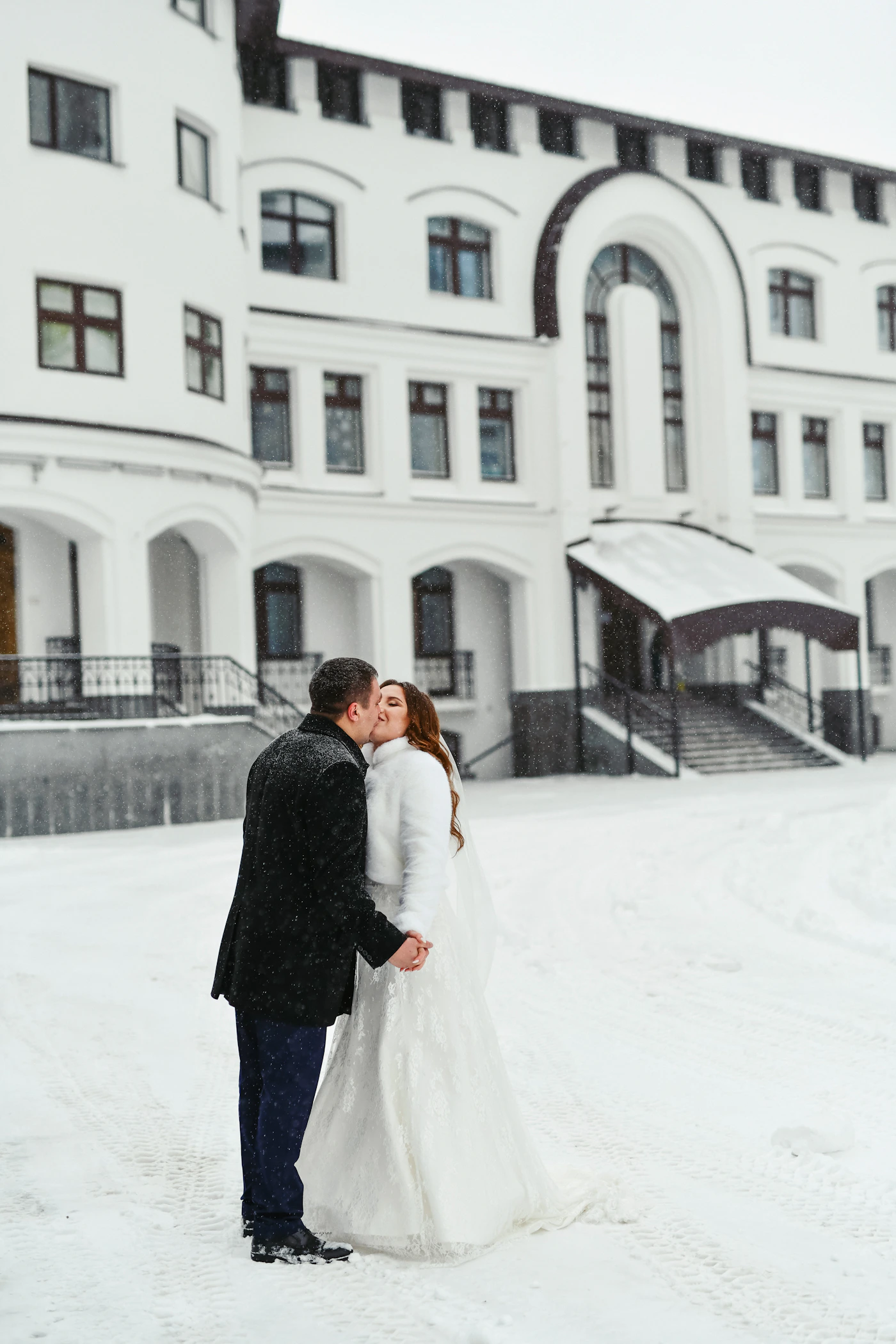 Bride and groom in wedding attire standing together in fresh snow with a grand white building behind them in winter