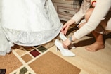 A person is helping another person put on white high-heeled shoes. The person being helped is wearing a white lace dress, suggesting a bridal outfit. They are indoors on a rug with a patterned design featuring various shades of beige, brown, and green colors. A chest of drawers is visible in the background.