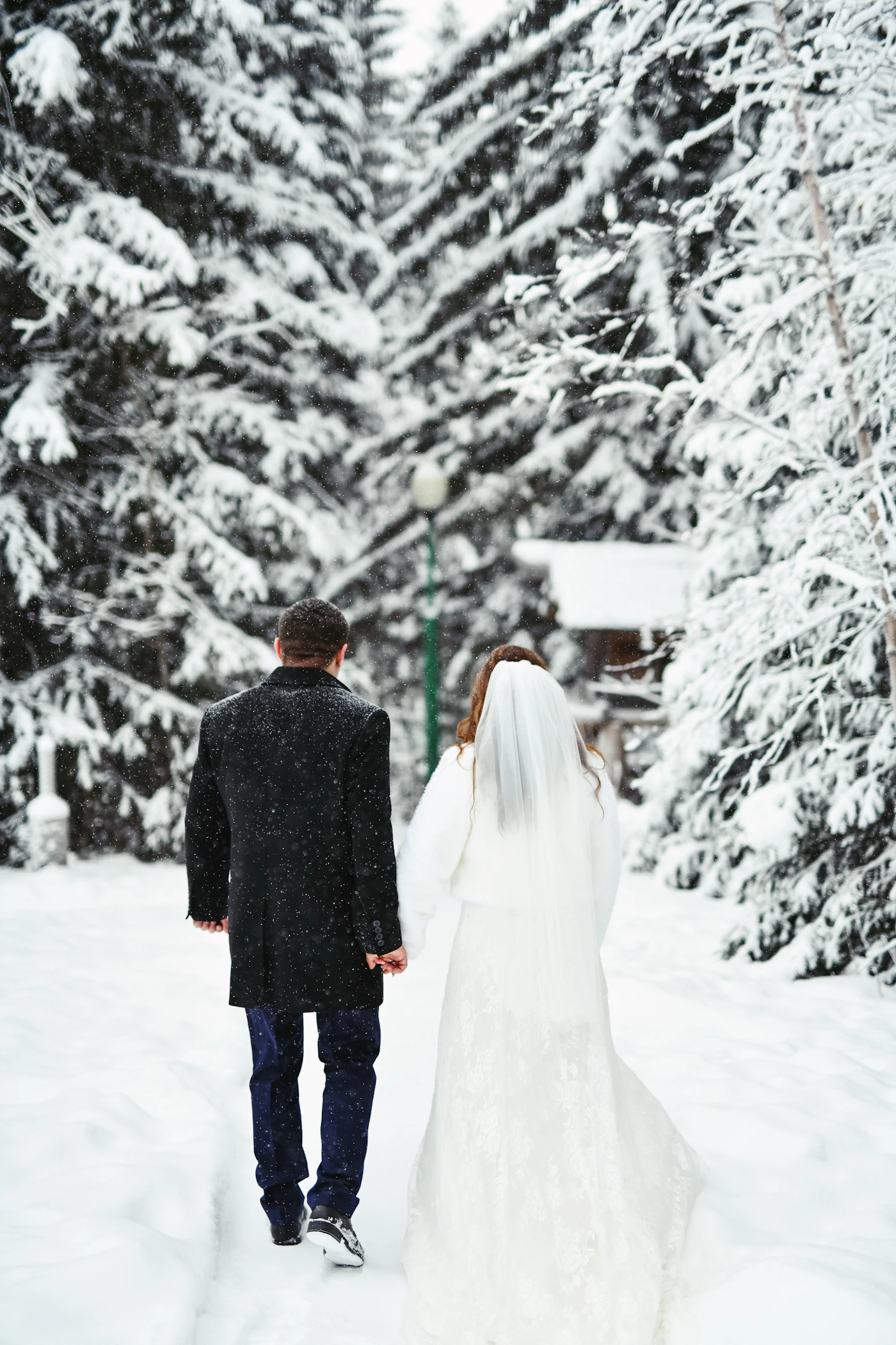Bride in white gown and groom in dark suit walking together through fresh snow during winter wedding with bare trees