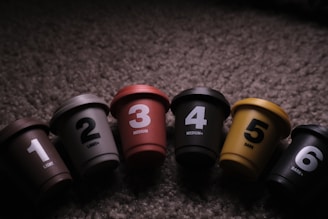 A series of six colorful coffee pod containers, each labeled with a number and corresponding roast level, are arranged on a textured surface. The containers display a gradient from light to dark roasts, with varying shades and colors.