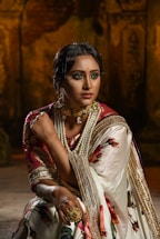 A woman is dressed in traditional attire, featuring a richly embroidered sari with floral patterns and golden accents. She is adorned with elaborate jewelry, including a choker necklace with pearls and a large ring. The background is dimly lit, adding a dramatic effect, emphasizing her poised and elegant demeanor.